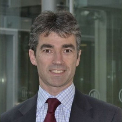 Headshot of Simon Buxton in a grey jacket, plaid shirt, and maroon tie