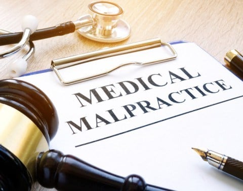 On a desk is a clipboard with top page titled MEDICAL MALPRACTICE, a gavel, fountain pen, and a stethoscope