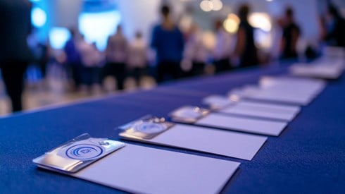 A conference registration desk with a row of nametags and notebooks, with conference attendees conversing in the background.