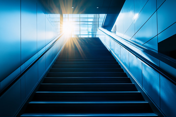 A staircase in a corporate office, leading upwards to bright sunlight shining through the lobby windows.