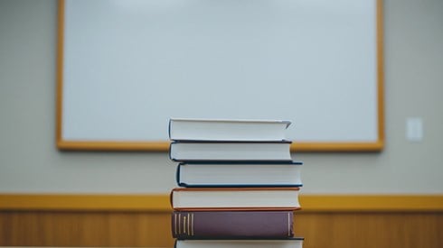 A stack of six textbooks with a blank whiteboard in the background.