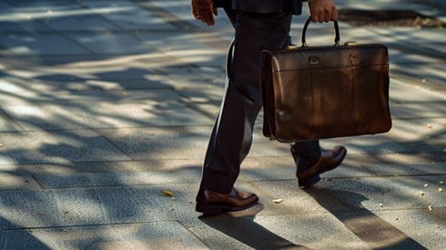 A businessman in a suit walking down a sidewalk while holding a brown leather briefcase.