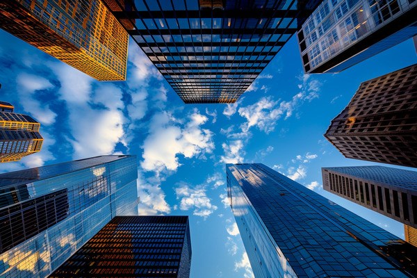 A collection of skyscrapers reaching towards the clouds in a blue sky, seen from street level looking upwards.