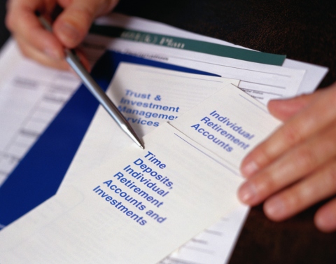 Two hands with a pen pointing to an Individual Retirement Account pamphlet with plan forms beneath it