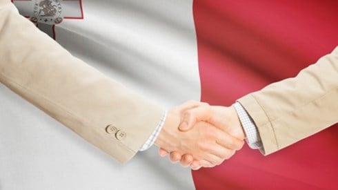 Two businessmen shake hands in front of the flag of Malta