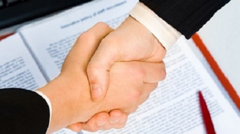 Two businessmen shaking hands over a contract in a red binder with a red pen and keyboard next to it