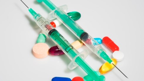 Two syringes with needles resting on top of pills forming the shape of a dollar sign