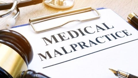 On a desk is a clipboard with top page titled MEDICAL MALPRACTICE, a gavel, fountain pen, and a stethoscope
