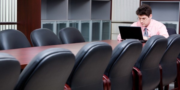 Man sitting at the end of a conference table in a large room working on a laptop