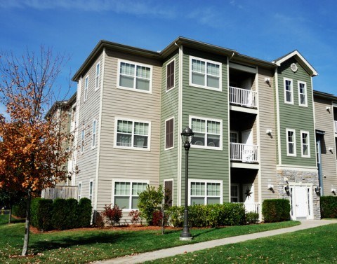 Green and taupe apartment complex building with green grass, a lamp post, and a tree with orange autumn leaves